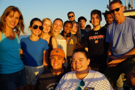 A group of smiling people gathered outdoors during sunset, with a boat railing and the sea in the background.