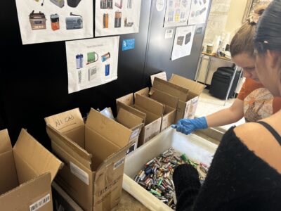 Two people sorting used batteries into labeled boxes at a recycling station, with battery type guides on the wall.