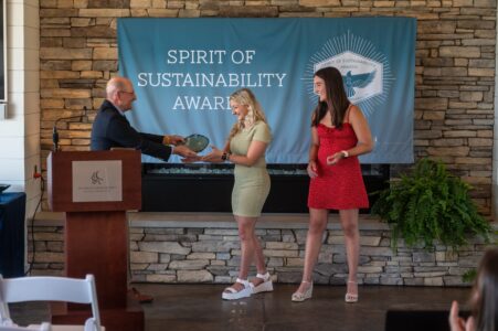 An award ceremony where a man hands a trophy to a woman, with another woman standing beside them. A "Spirit of Sustainability Awards" banner is in the background.