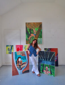 Woman posing with her paintings in a bright art studio.
