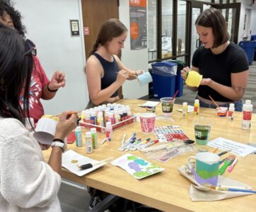 a group of students painting mugs at a table with craft supplies. 