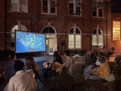 a group of people gathered in an outside courtyard looking at a projector screening of "Kiss the Ground"