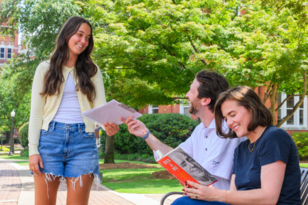A young person stands smiling while handing a clipboard to two seated individuals who are also smiling and engaged in a pleasant outdoor interaction.
